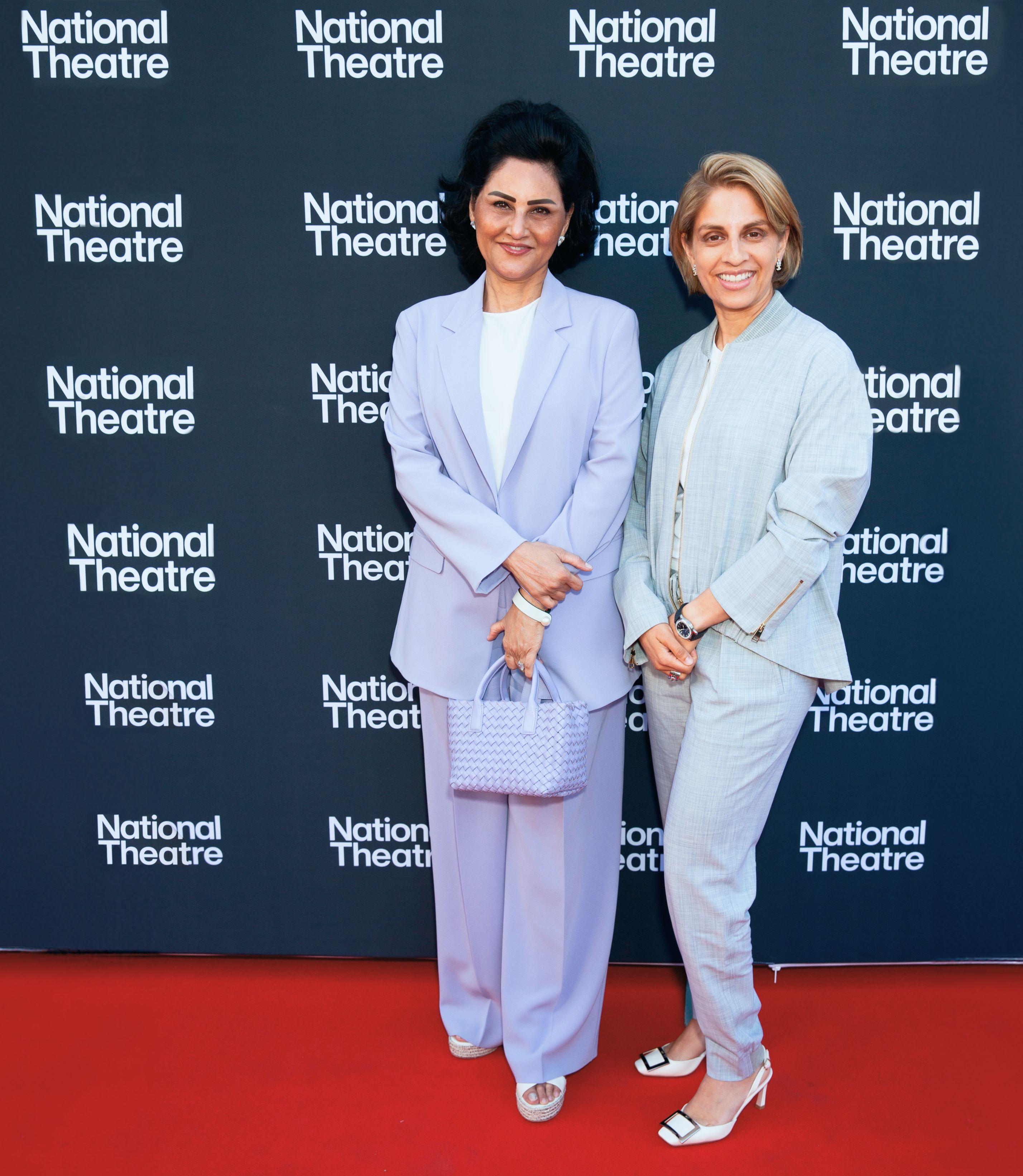 Two women of South Asian ethnicity stand on a red carpet. One has black, shoulder length hair and wears a lilac suit. The other has a blonde bob and wears a light gray suit.